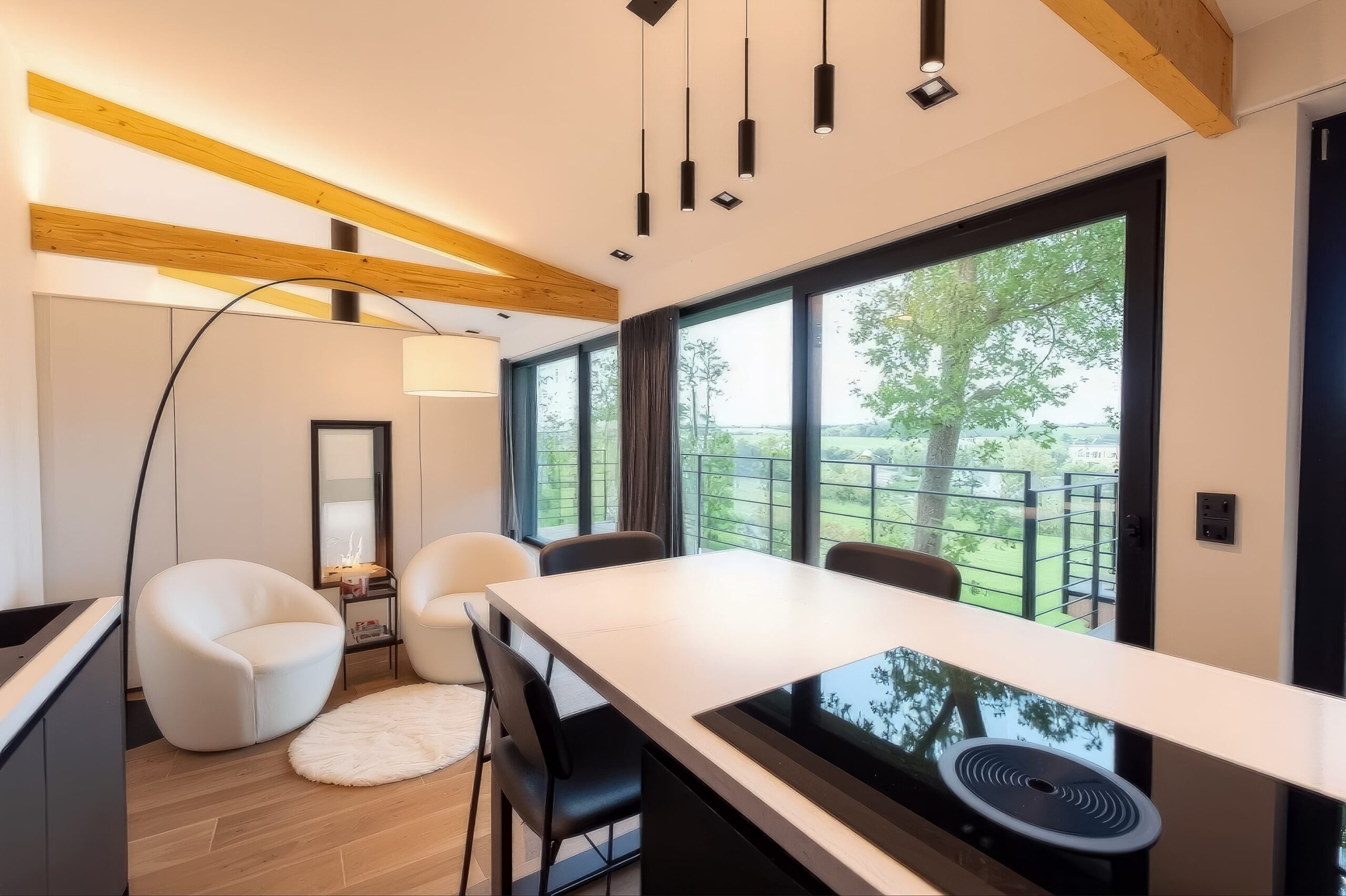 Vue panoramique depuis l'îlot de cuisine sur la nature verdoyante de Gesves, avec espace repas convivial et coin détente avec fauteuils blancs.