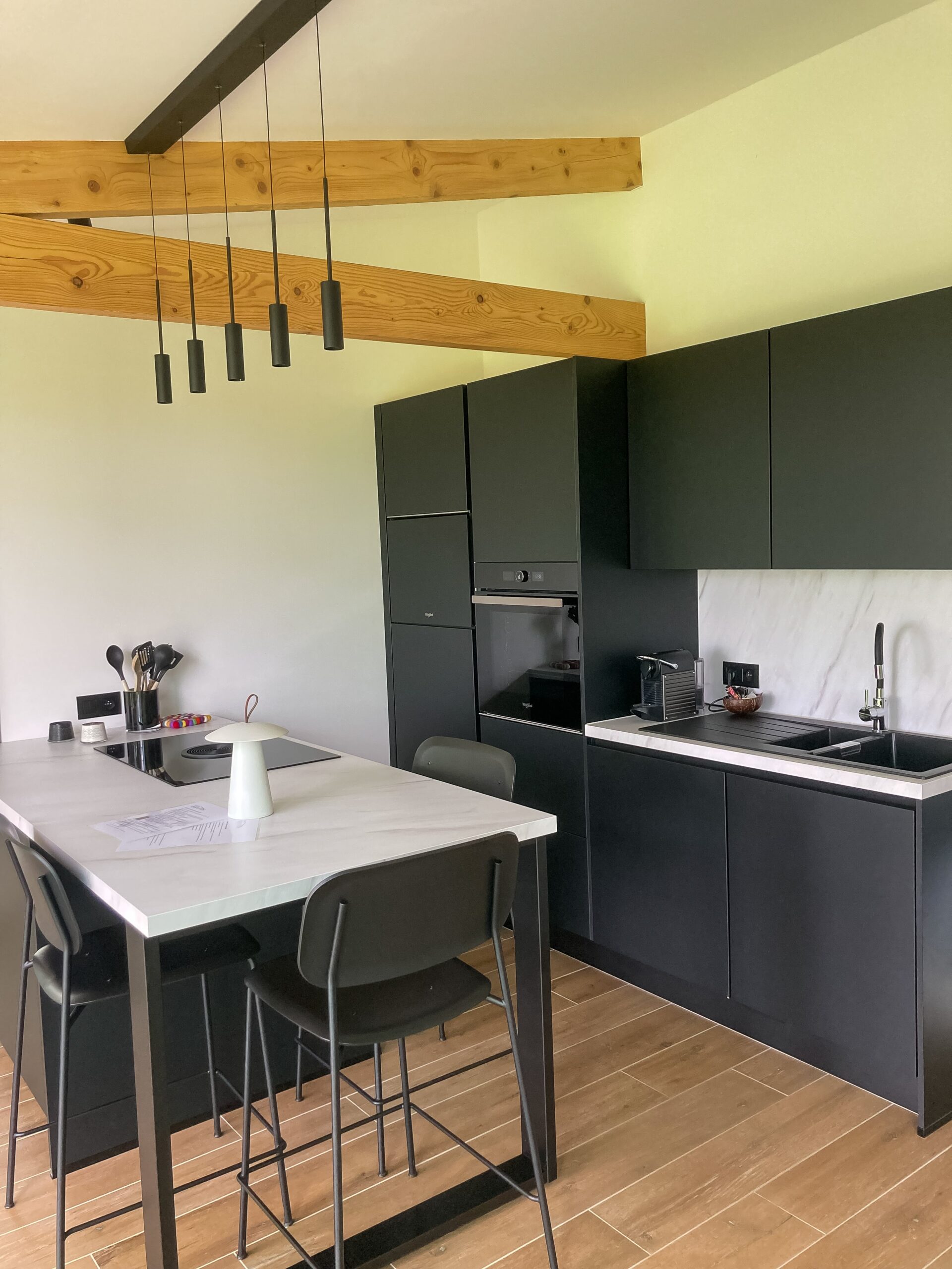 Vue d'ensemble de la cuisine équipée noire : îlot central avec coin repas, suspensions luminaires noires et intégration sous poutres en bois.