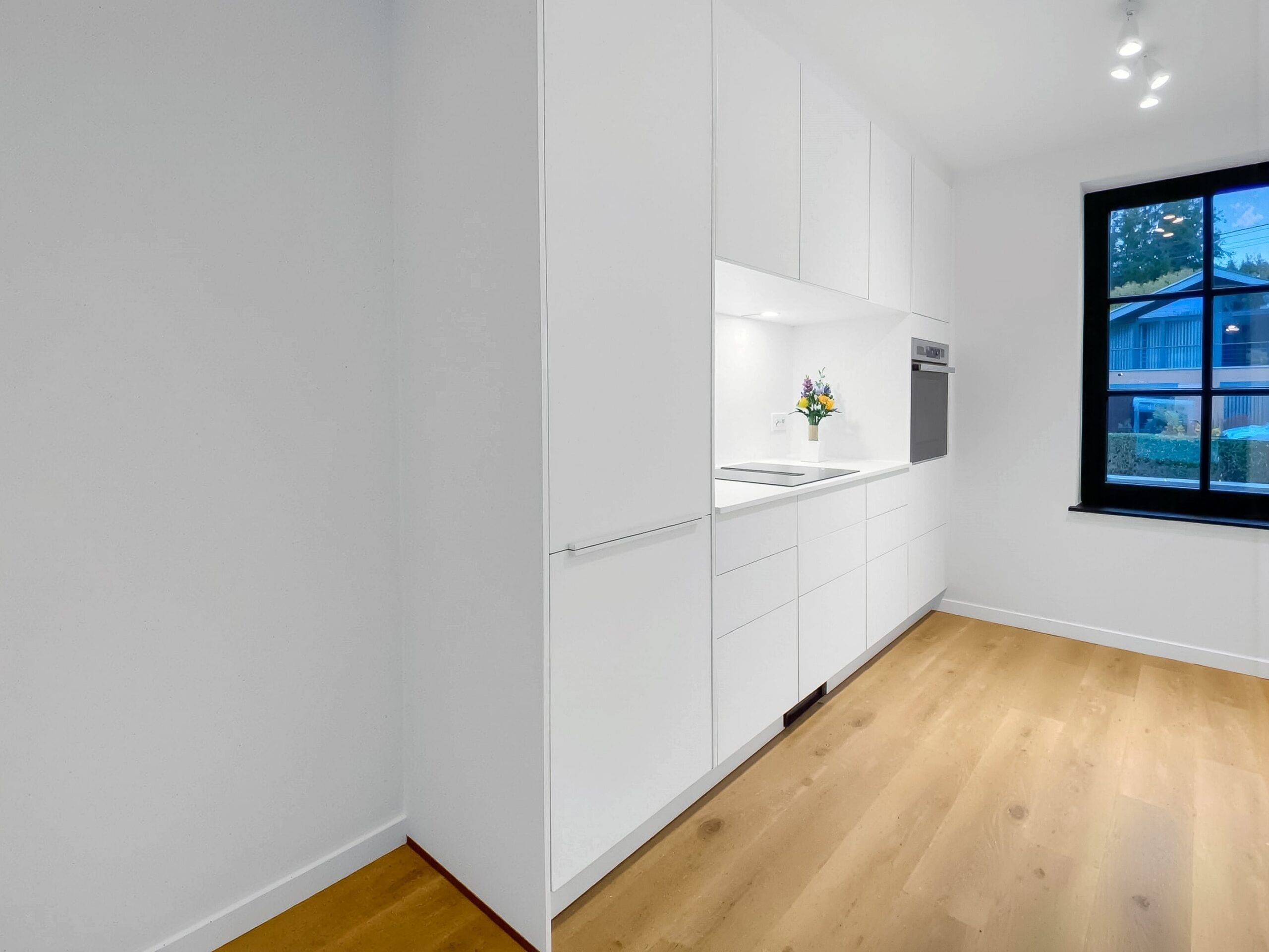 Cuisine minimaliste blanche K-Lita à Waremme avec rangements toute hauteur, plan de travail épuré et sol en parquet clair dans une pièce lumineuse.