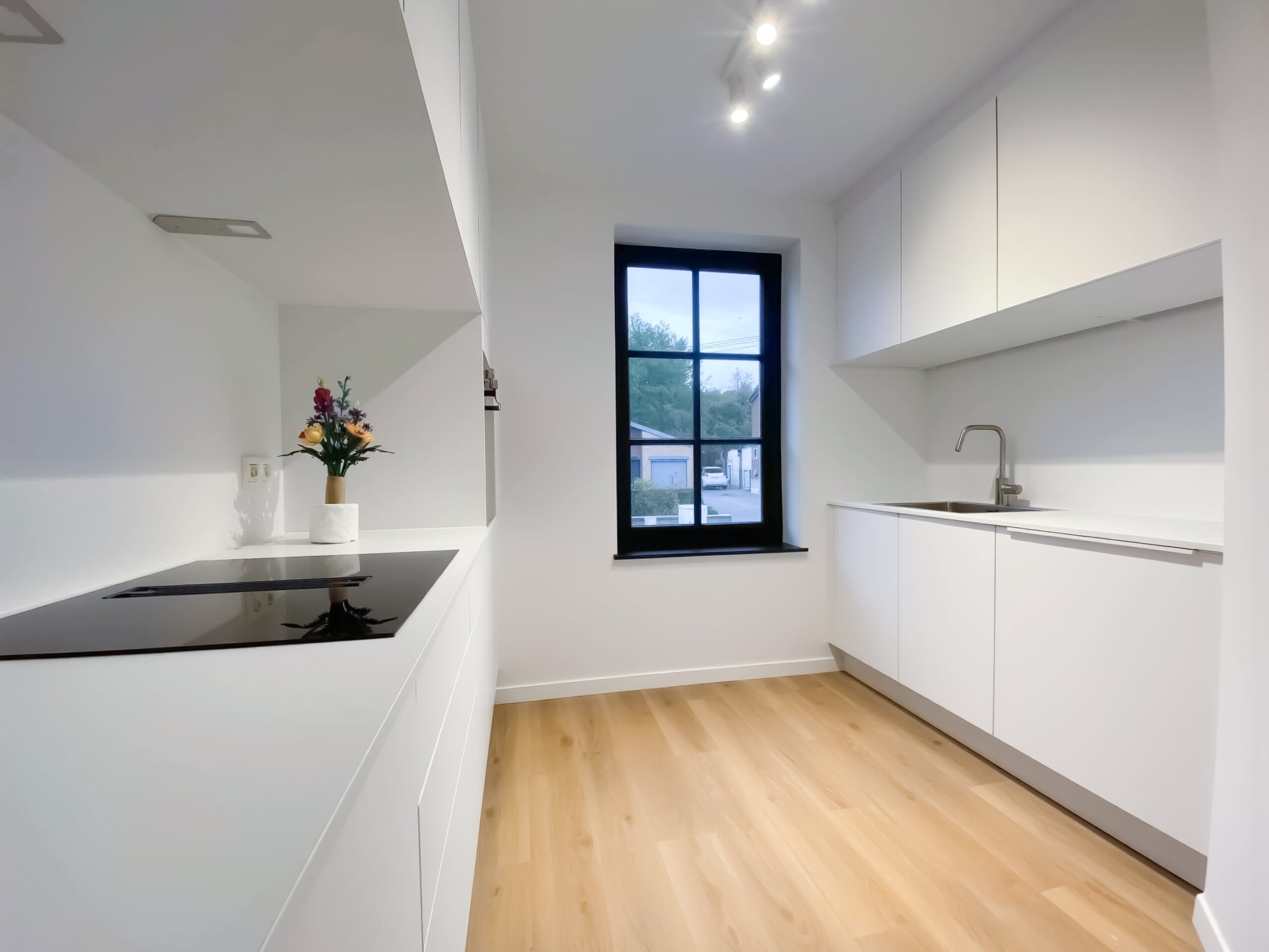 Vue de face d'une cuisine équipée blanche moderne : évier inox, taque de cuisson à induction et fenêtre à châssis noir offrant une vue sur l'extérieur à Waremme.